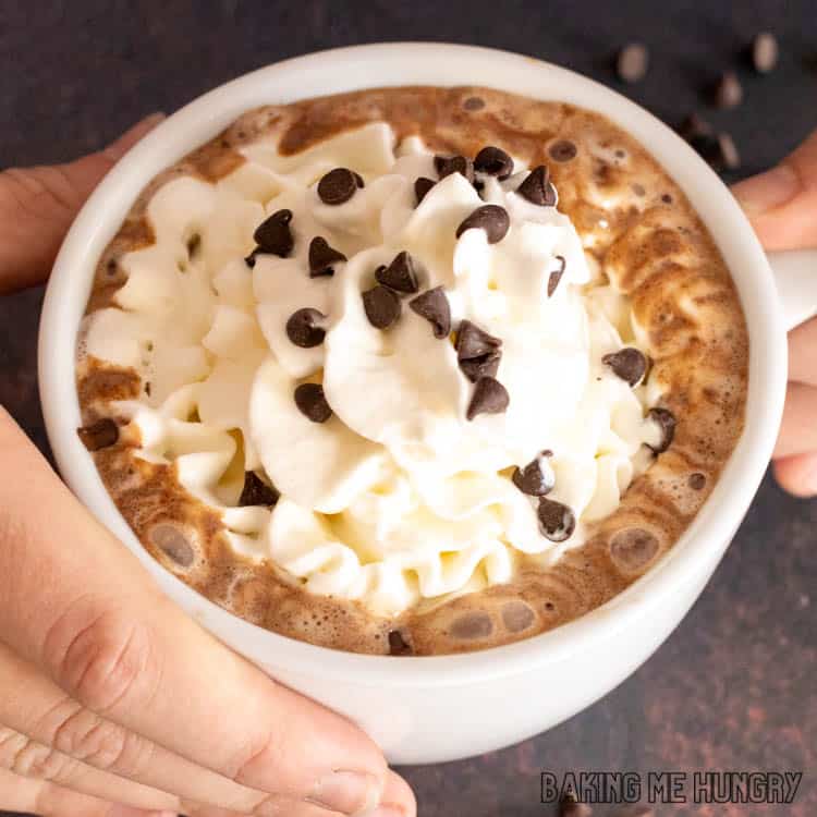 overhead shot of hands holding mug with chocolate chips hot chocolate in mug topped with whipped cream and mini chocolate chips