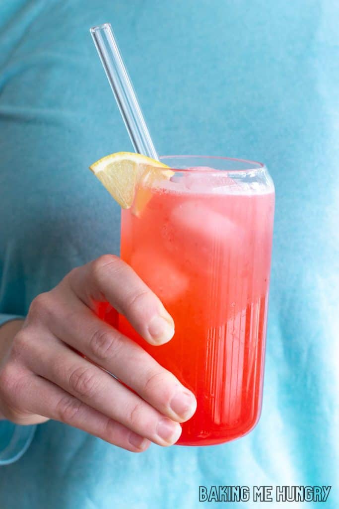 raspberry lemonade recipe in a tall glass with a lemon slice as garnish held by a hand in front of a blue shirt
