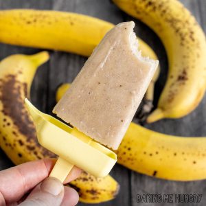 creamy banana popsicles recipe shown as a hand holding up a pop missing a bite with bananas behind